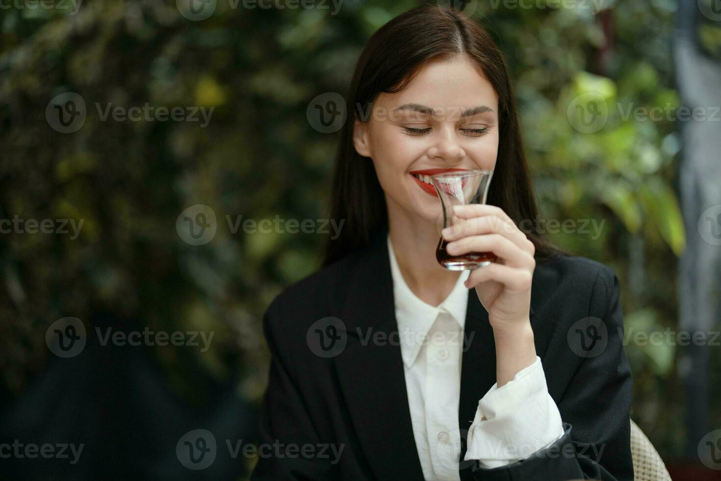 kvinna med röd mun leende med tänder drycker te i en Kafé från en turkiska glas råna i en vit skjorta och svart mode jacka på de gata, sommar resa, semester i de stad foto