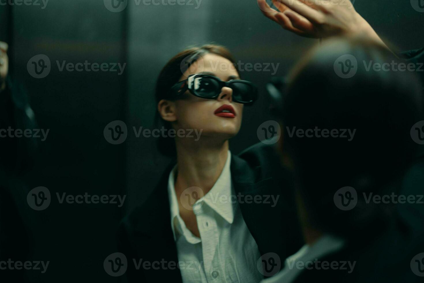 eleganta kvinna i en svart jacka och solglasögon Framställ i ett hiss hiss nära de spegel, mode modell, mörk filmiska ljus och Färg matris, glamour årgång foto