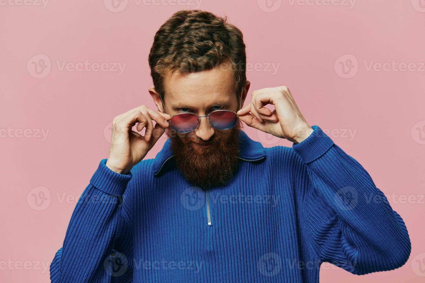 porträtt av en rödhårig man bär solglasögon leende och dans, lyssnande till musik på en rosa bakgrund. hipster med en skägg, lycka finger pekande. foto