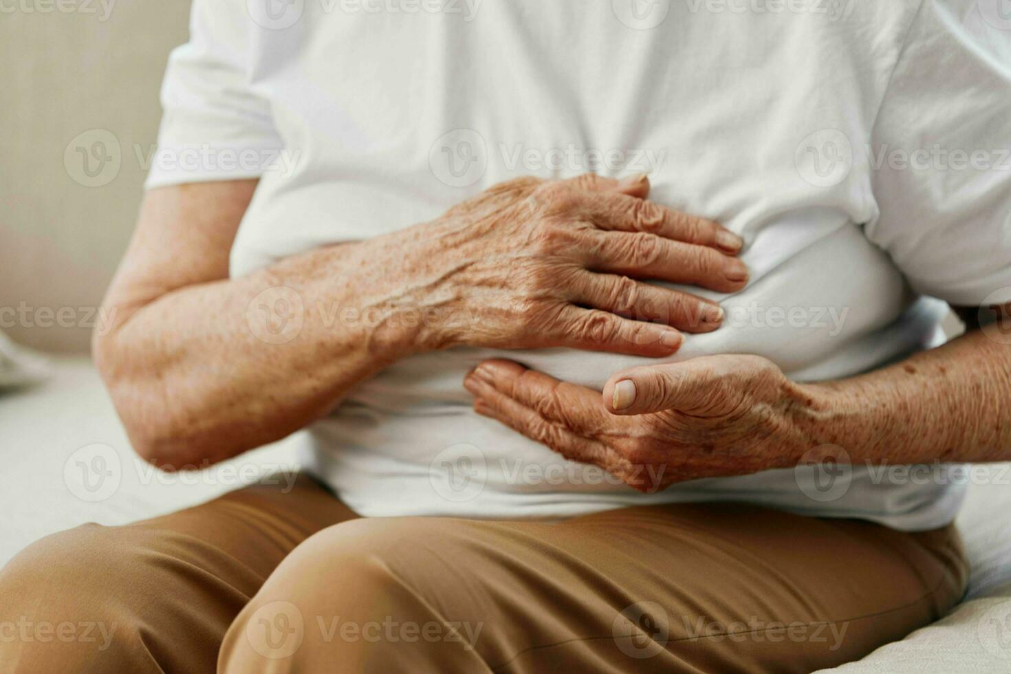 äldre kvinna svår bröst smärta Sammanträde på de soffa, hälsa problem i gammal ålder, fattig kvalitet av liv. mormor med grå hår innehar henne bröst med henne händer, kvinnors hälsa, bröst cancer. foto