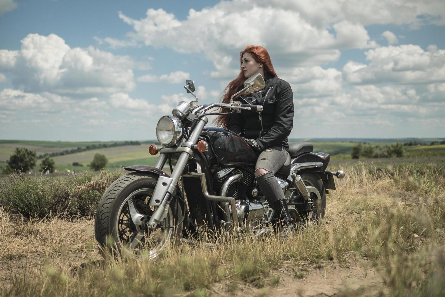 den rödhåriga cykelflickan sitter på ett motorcykelfält av äng och moln foto