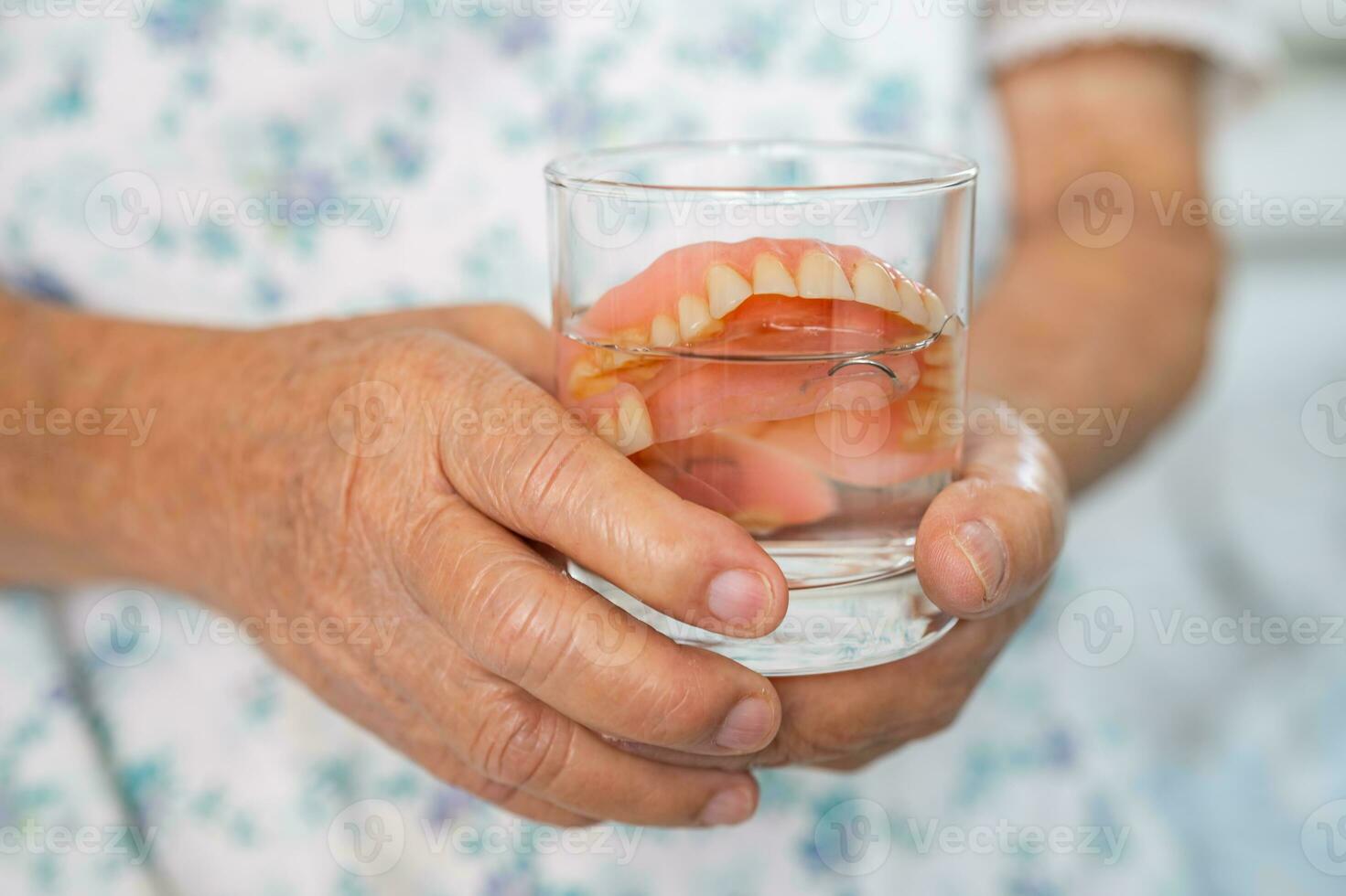 asiatisk senior kvinna patient innehav och tvättning tandprotes i vatten rengöringsmedel glas för Bra tugga. foto