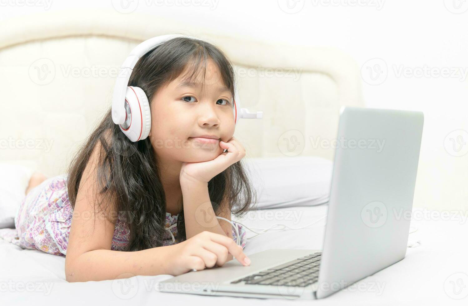 Lycklig söt flicka liggande och bär hörlurar till lära sig uppkopplad från en bärbar dator foto