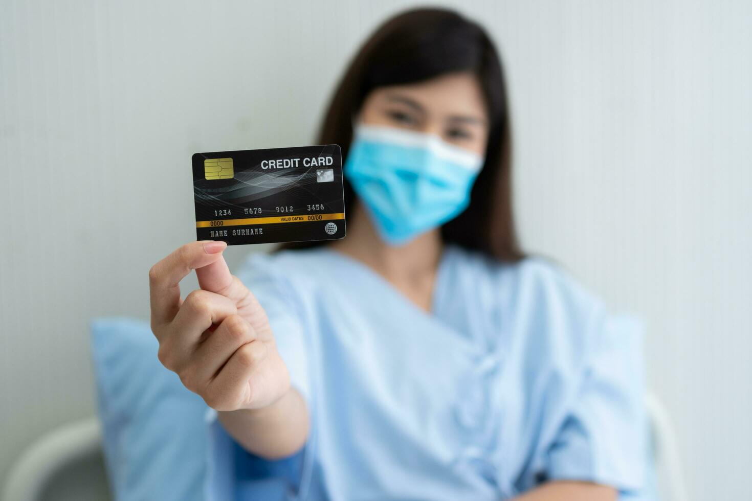 glad asiatisk kvinna som bär en medicinsk mask och håller mock up kreditkort försäkringskort och ok tecken i en sjukhussäng. försäkring av bank, betalning medicinsk behandling koncept foto