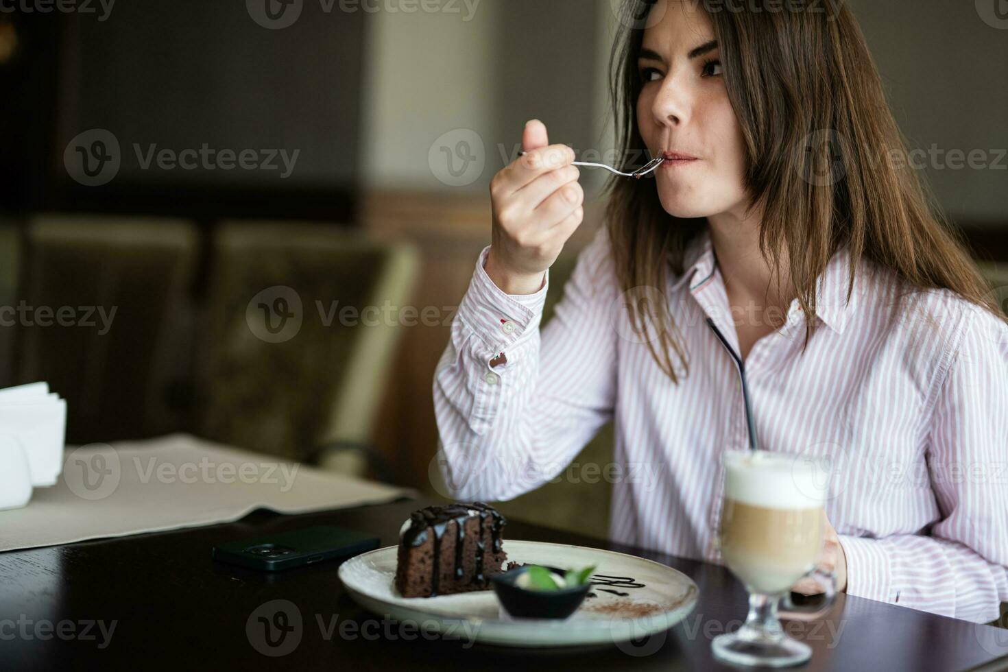 ung skön brunett kvinna sitta i kaffe affär Kafé restaurang inomhus och äta choklad tomte efterrätt kaka, gaffel nära de mun. foto