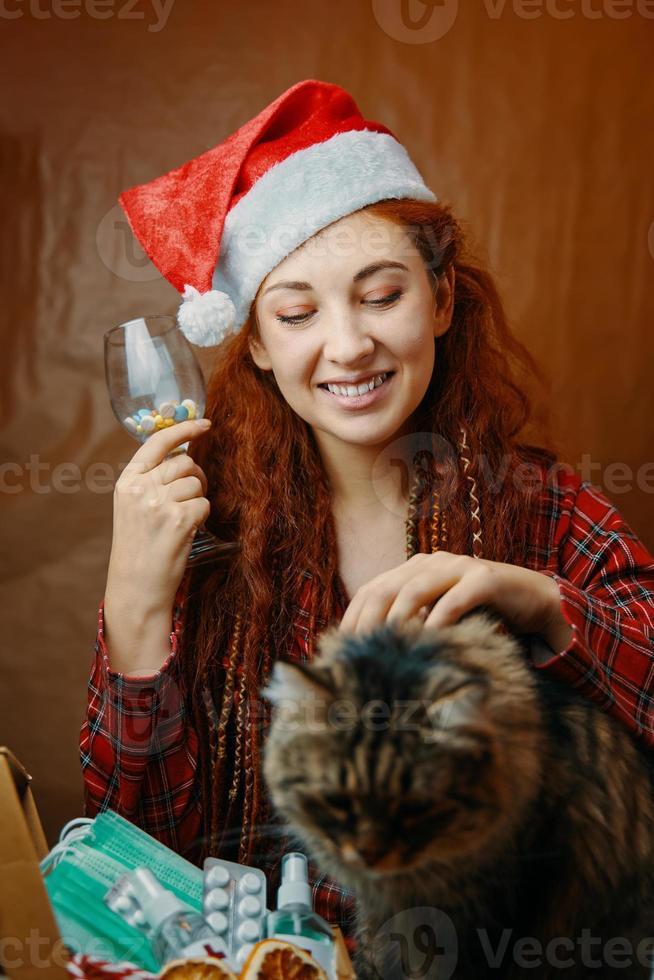 rolig tjej i santa hatt håll glas piller och stroke katt foto