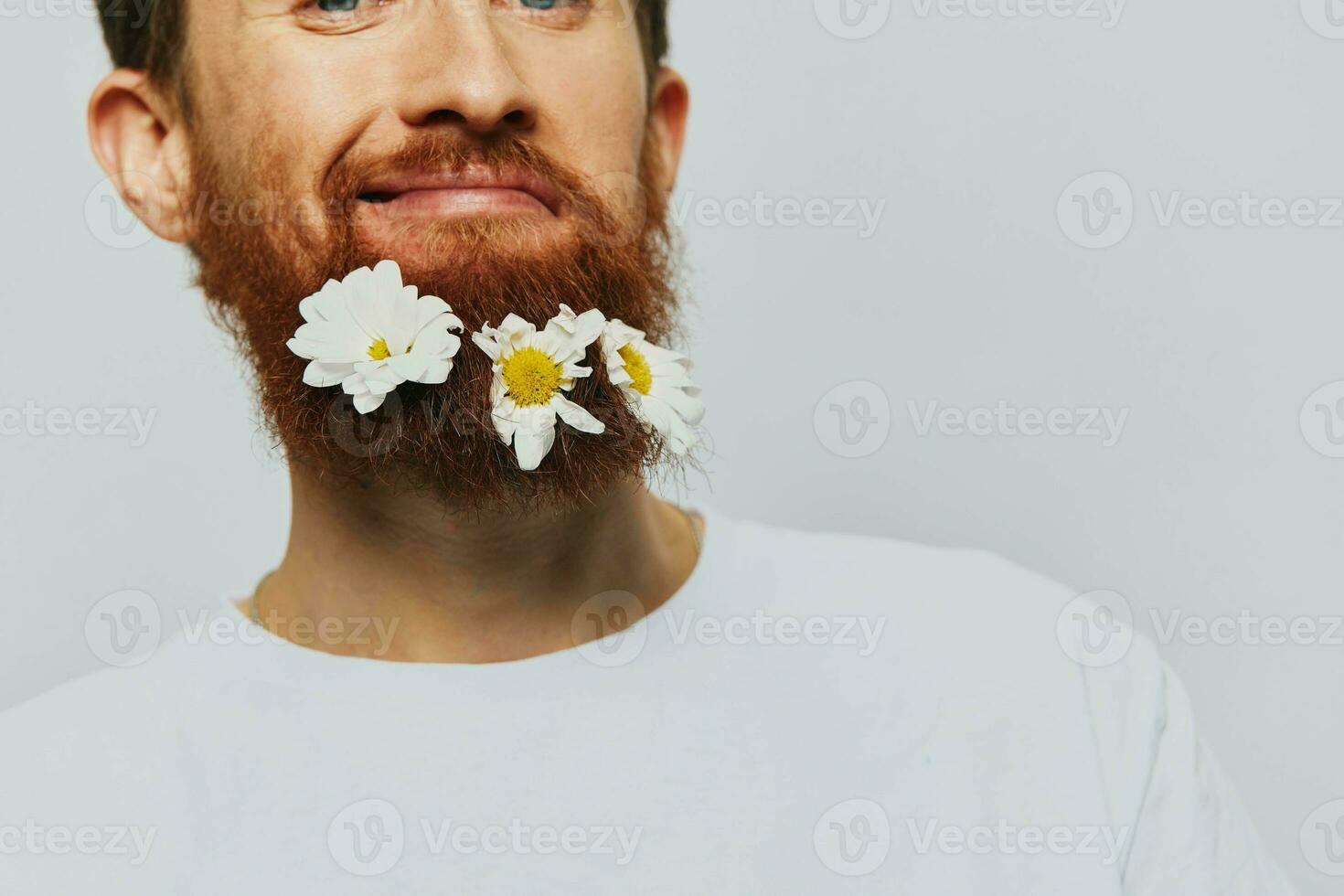 porträtt av en rolig man i en vit t-shirt med blommor daisy i hans skägg på en vit isolerat bakgrund, kopia plats. Semester begrepp och grattis. foto