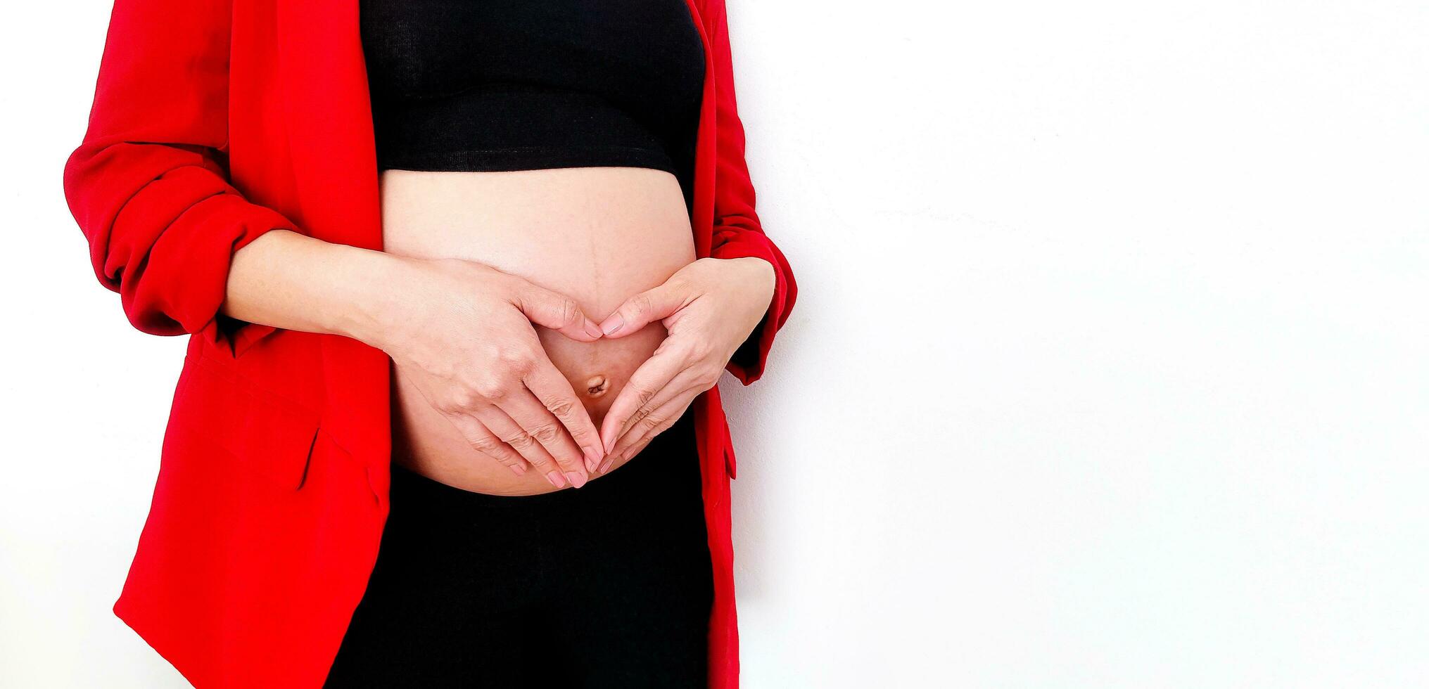 stänga upp gravid kvinna i röd kostym formning en hjärtformad med henne händer över henne mage eller mage isolerat på vit bakgrund och kopia Plats med klippning väg. graviditet 7-9 månader, moderskap foto