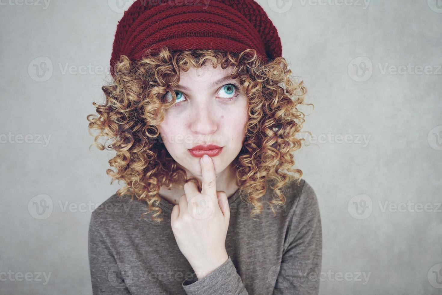 nära porträtt av en vacker och ung rolig tankeväckande kvinna med blå ögon och lockigt blont hår som tänker och bär en röd ullmössa foto