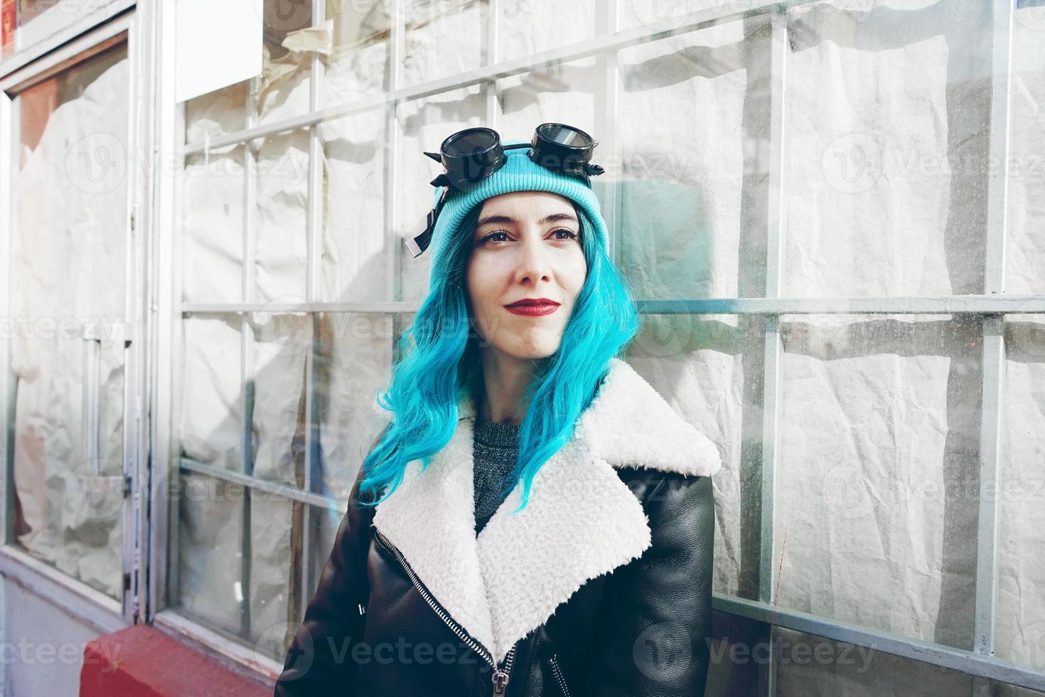 porträtt av en punk eller gotisk ung kvinna med blått färgat hår och bär svarta steampunk-glasögon och blå ullmössa i en stadsgata utomhus foto