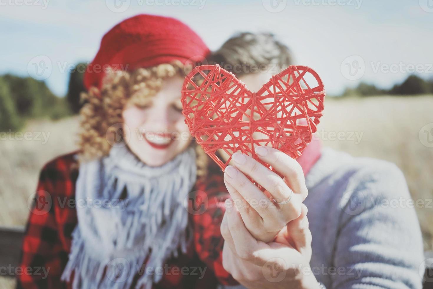 härlig närbild av ett vackert rött hjärta som ryms av ett romantiskt ungt par i kärlek i en trevlig bokeh som bakgrund utomhus. det påminner kärlek eller hälsovård foto