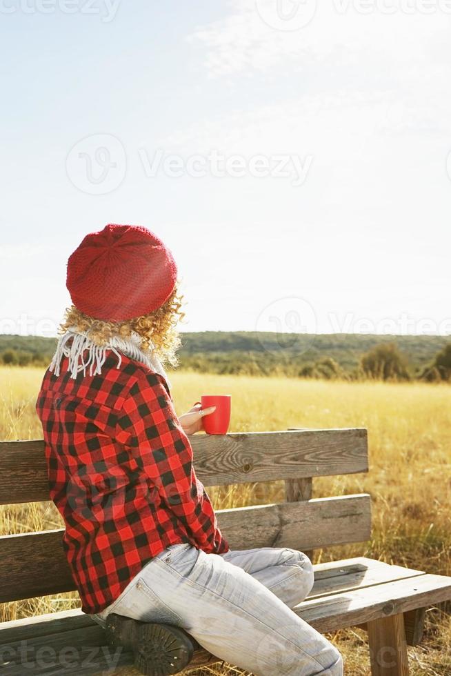 en ung kvinna bakifrån i röd rutig skjorta med ulllock och halsduk som tar en kopp te eller kaffe medan hon solar sittande i en träbänk i ett gult fält med bakgrundsbelysning från höstsolen foto