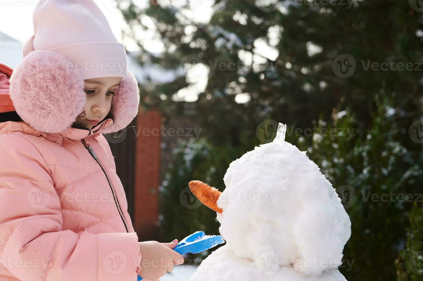 närbild porträtt härlig liten barn flicka i rosa ner jacka och fluffig hörselkåpor, byggnad snögubbe i snöig bakgård foto