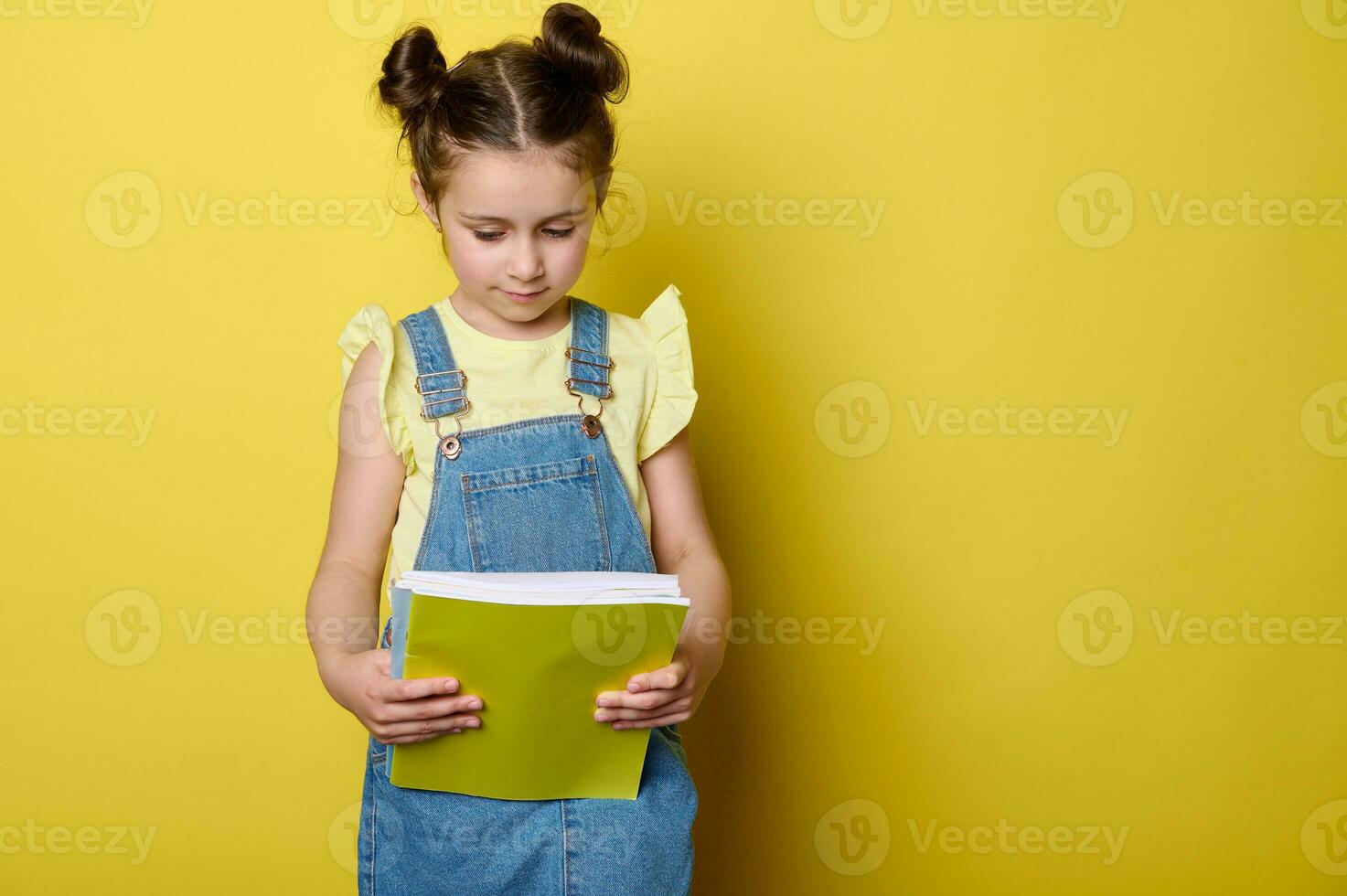 porträtt av söt barn flicka i gul t-shirt och blå denim klänning overall, innehav lärobok isolerat studio bakgrund foto