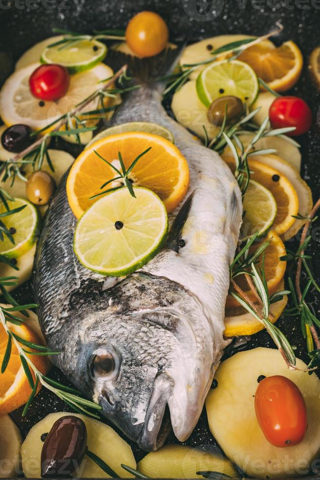 havsgyltad braxfisk i bakplåten redo att bakas med potatis, rosmarin, citron, apelsin, oliver, tomater, lök och lime. färsk orata, doradefiskberedning foto