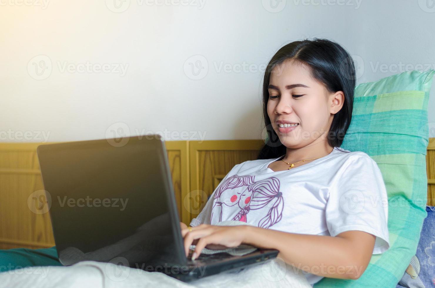 glad avslappnad vacker asiatisk kvinna som arbetar på en bärbar dator anteckningsbok sova på sängen i huset. foto