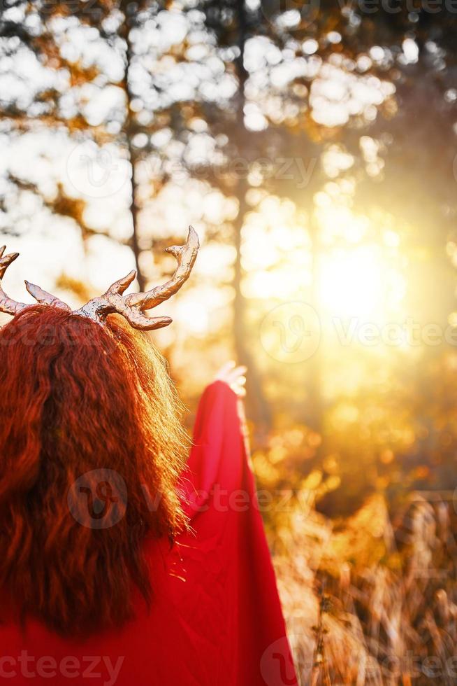 kvinna i lång röd klänning med hjorthorn i höstskog försöker röra vid en solnedgång foto