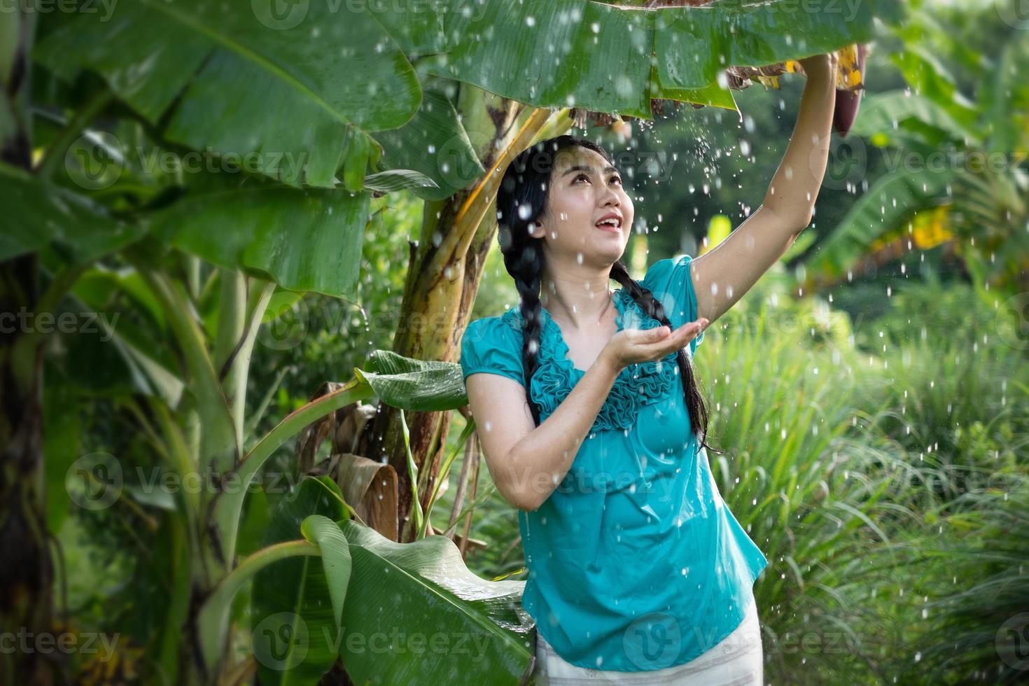 porträtt av en ung asiatisk kvinna med svart hår som håller ett bananblad i regnet på den gröna trädgårdsbakgrunden foto