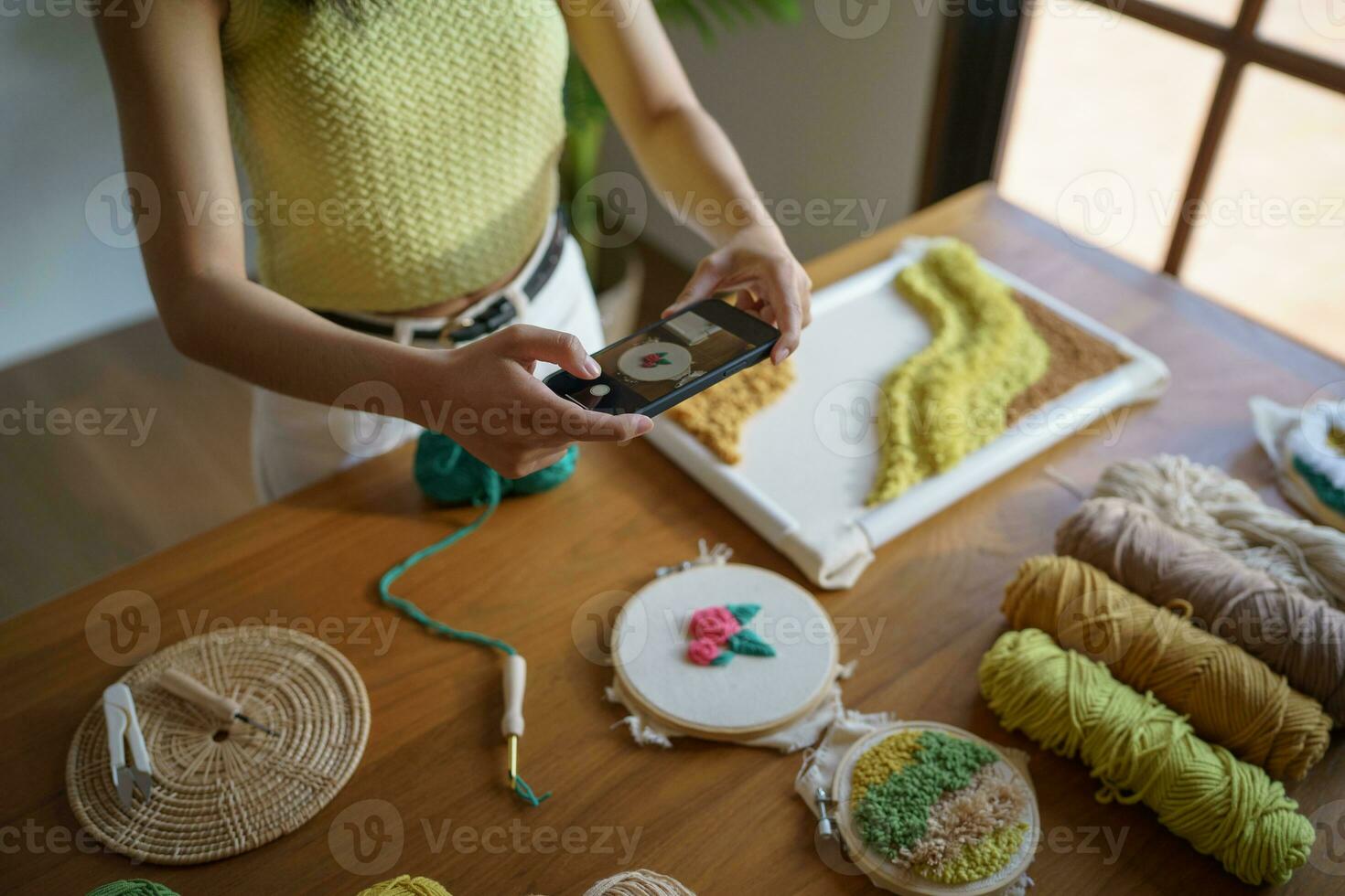 asiatisk kvinna tar smartphone Foto stansa nål. telefon inlägg på social nät i studio verkstad. designer arbetsplats handgjort hantverk projekt diy broderi