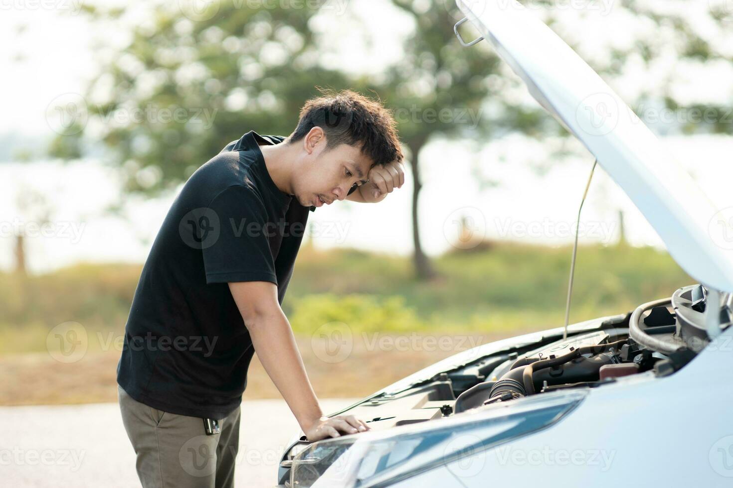 orolig asiatisk ung man står i främre av bil kontroll tillstånd efter en bruten bil. bruten bil ner på de väg. nödsituation service har bruten bil. foto