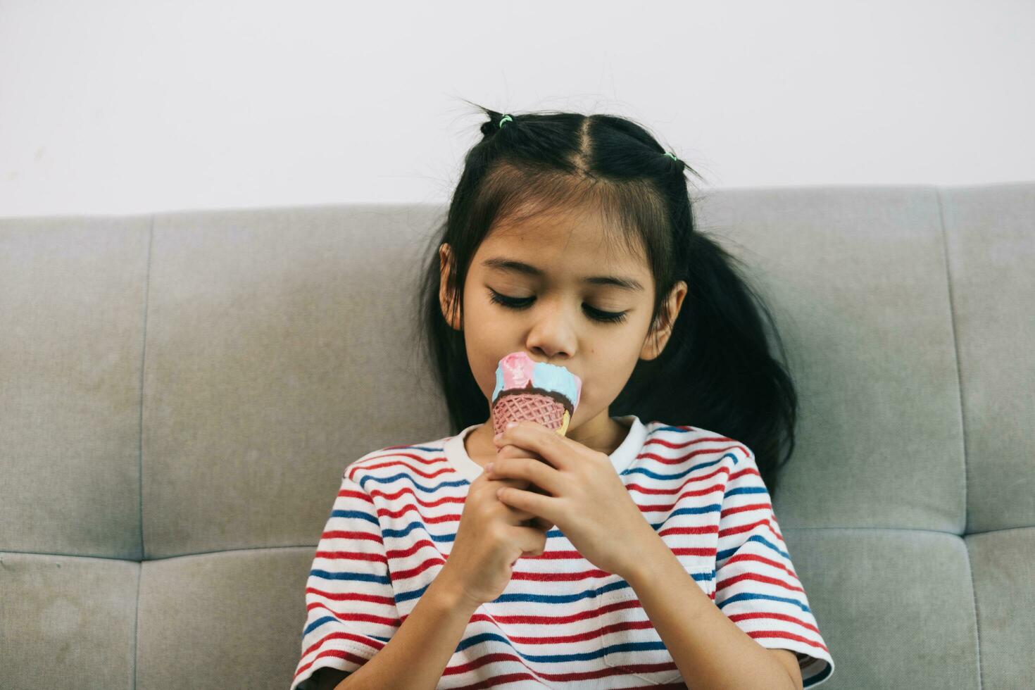 liten asiatisk flicka äter is grädde på soffa i levande rum. foto