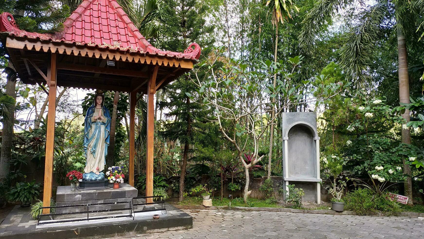 staty av de jungfrulig mary under en tak med träd i de bakgrund, i de jatiningsih maria grotta, jogjakarta, var katoliker be, ge tack och Skicka in begäranden till Gud foto