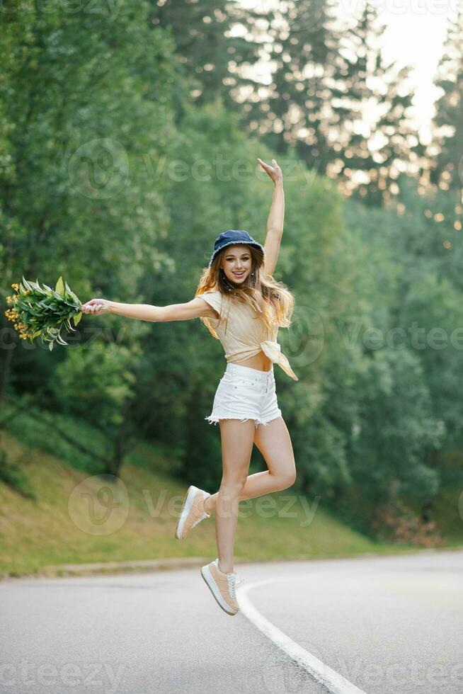 en Lycklig flicka i vit shorts och en gul blus hoppar upp med en bukett av blommor, stående på de väg nära de skog foto