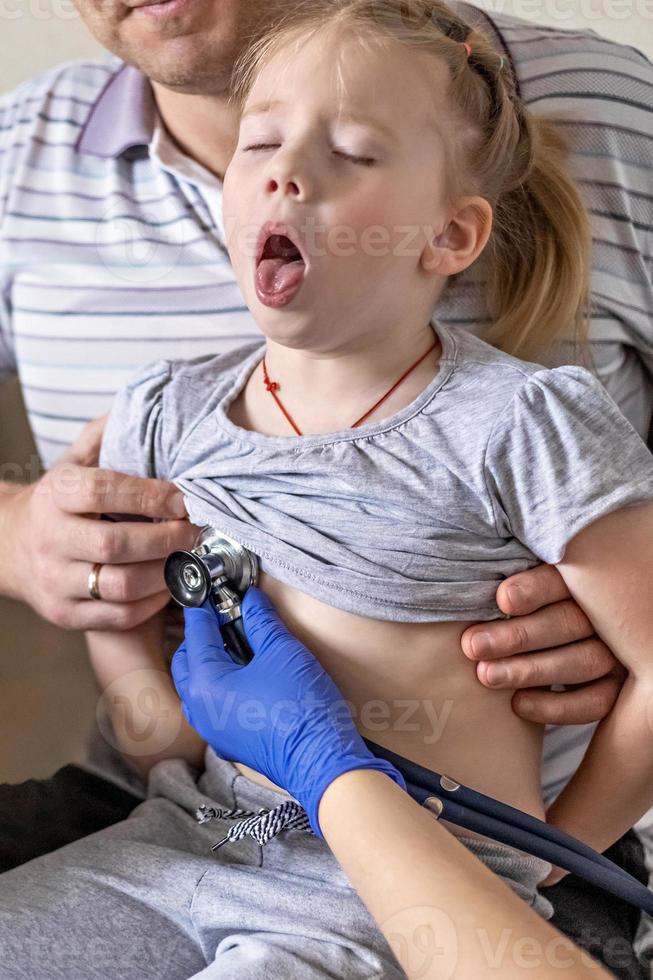 liten flicka i armarna på sin far på läkarmottagningen på kliniken. läkaren undersöker barnet, lyssnar på lungorna med ett fonendoskop. behandling och förebyggande av luftvägsinfektioner. foto