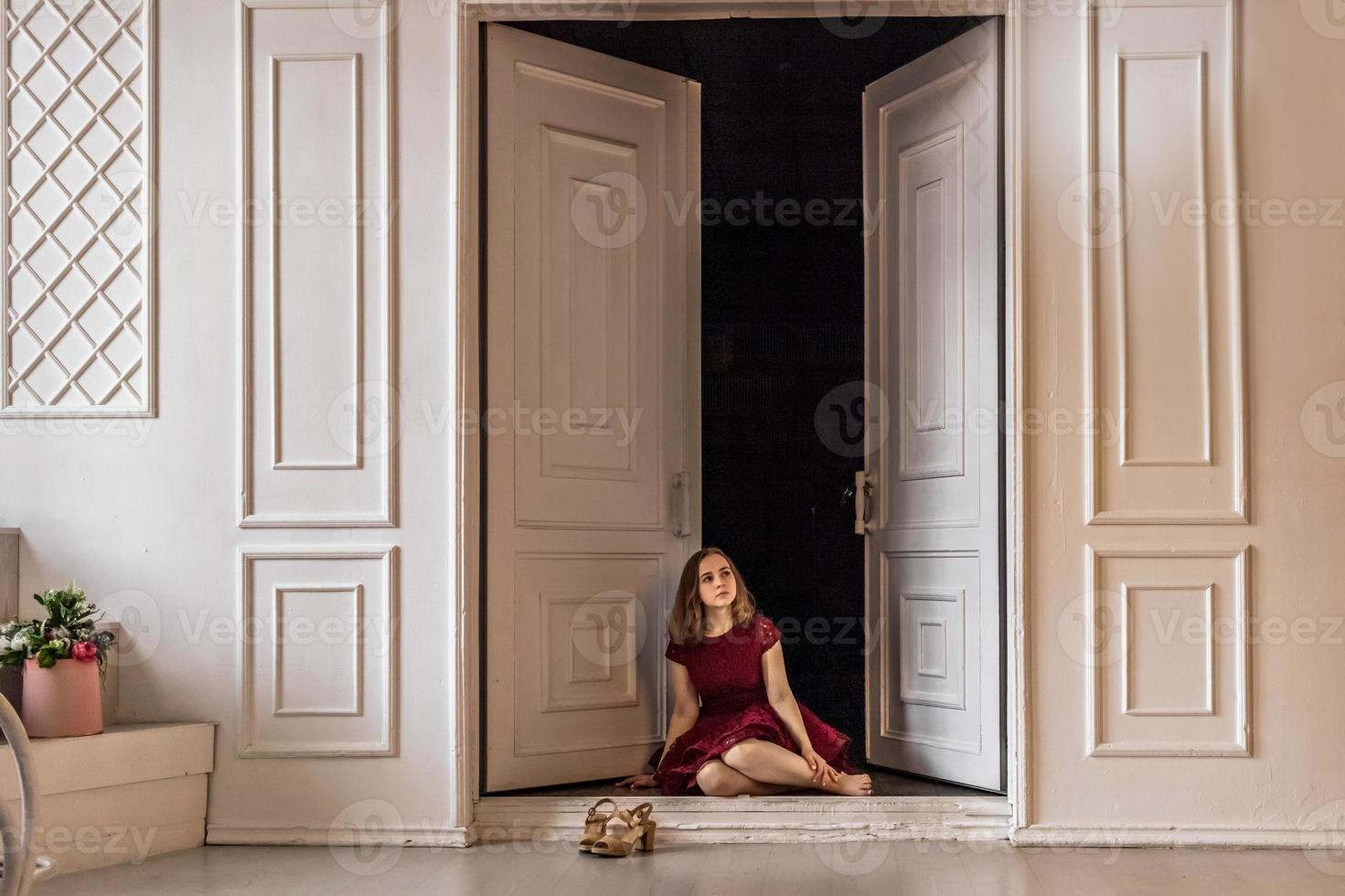 en söt snygg ung flicka i en elegant vinröd klänning sitter i dörren till sitt rum. tonåring. examen i skolan, college foto