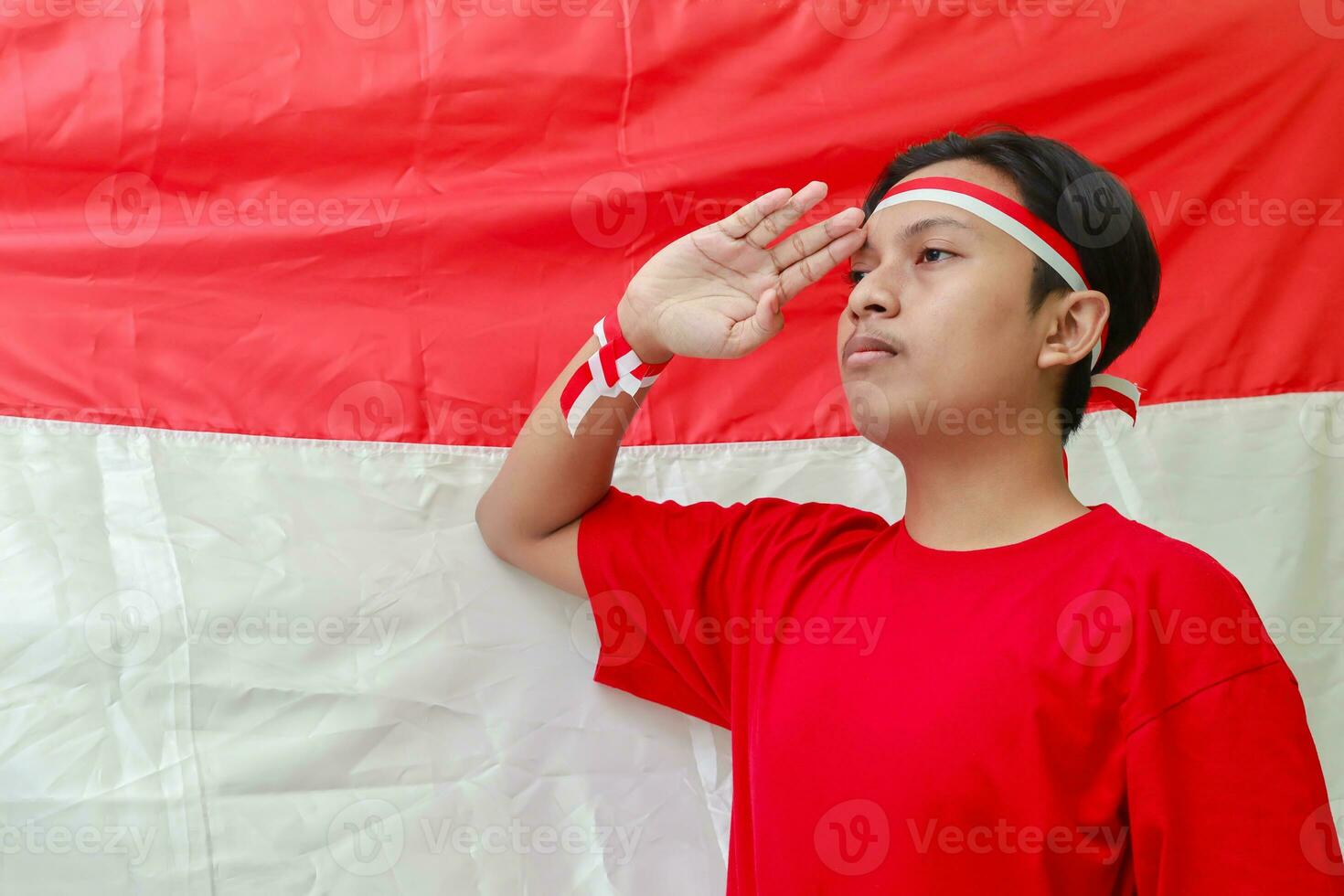 porträtt av attraktiv asiatisk man i röd skjorta med röd och vit band på huvud, ger honnör utgör med hand i främre av indonesiska flagga foto