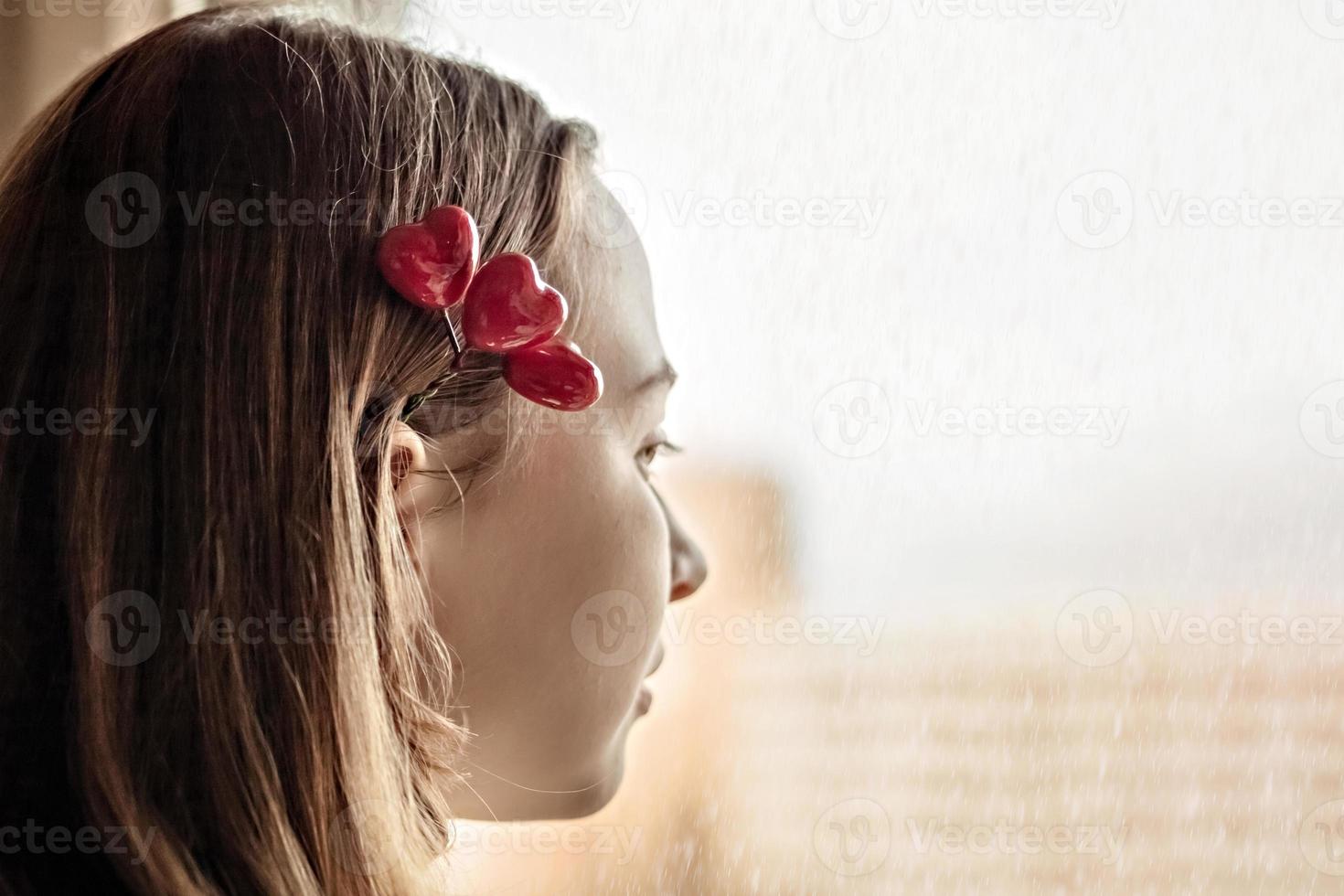 porträtt av en vacker sorglig tjej som ser ut genom fönstret med hopp och förväntan. hårnål med hjärtan. första kärleken på alla hjärtans dag foto