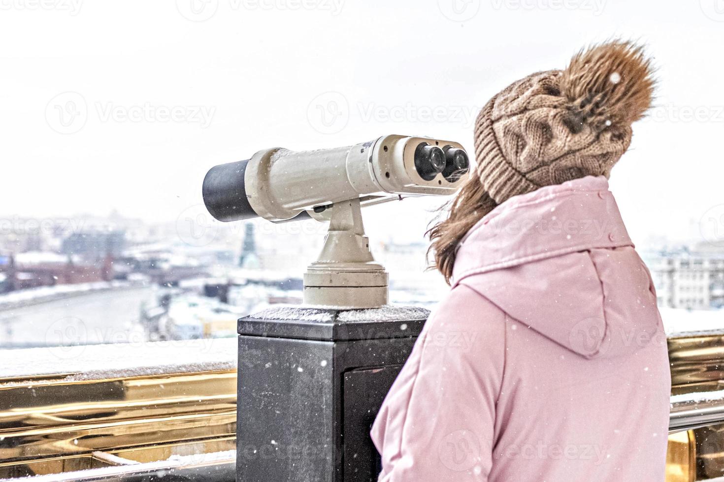en ung flicka tittar genom myntdrivna kikare på observationsdäcket med utsikt över staden från en höjd vid solnedgången. vinter, snöfall foto