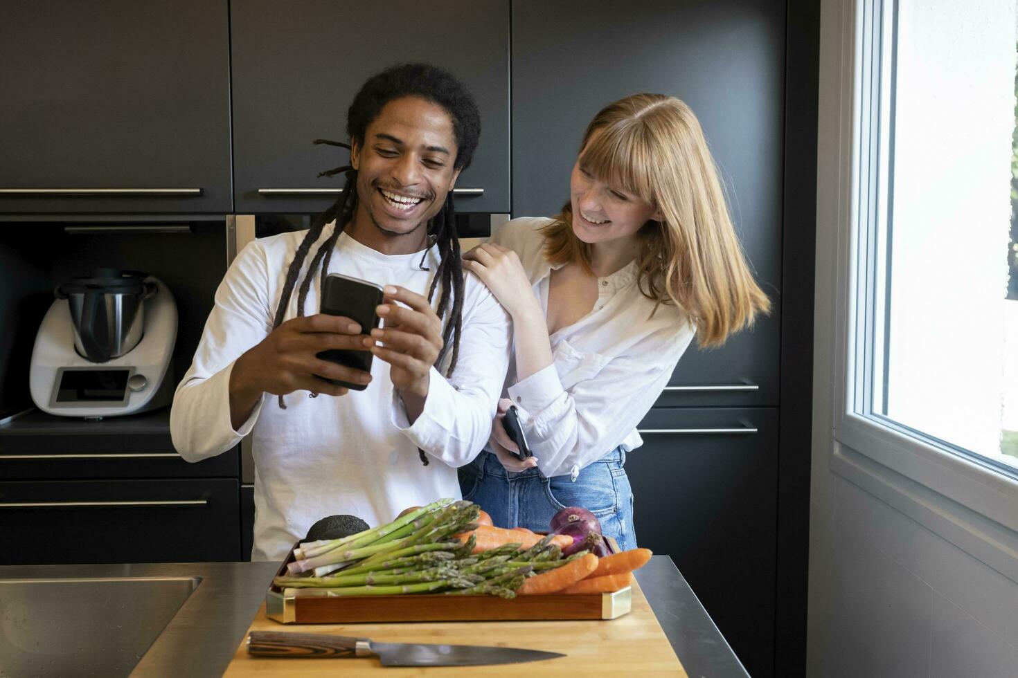 ung par av annorlunda etnisk grupper med mobil telefon i en kök med vegetabiliska styrelse. begrepp av friska levande. foto