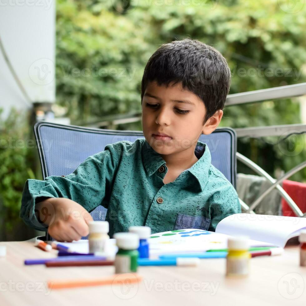 smart indisk liten pojke prestera tumme målning med annorlunda färgrik vatten Färg utrustning under de sommar semester, söt indisk unge håller på med färgrik tumme målning teckning på trä- tabell foto