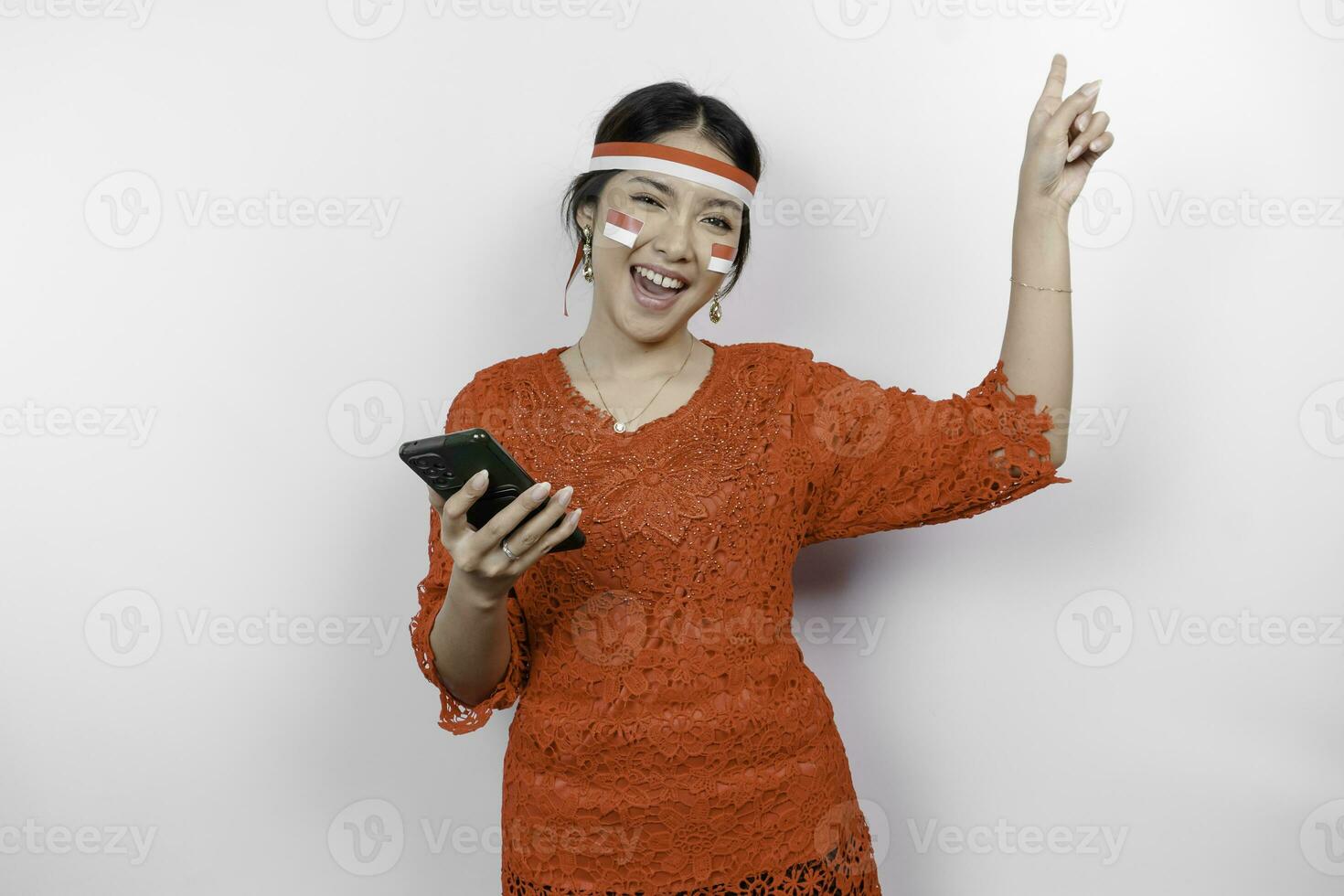 en Lycklig asiatisk kvinna bär röd kebaya och pannband, innehav henne telefon, och pekande kopia Plats på topp av henne, isolerat förbi vit bakgrund. Indonesiens oberoende dag foto