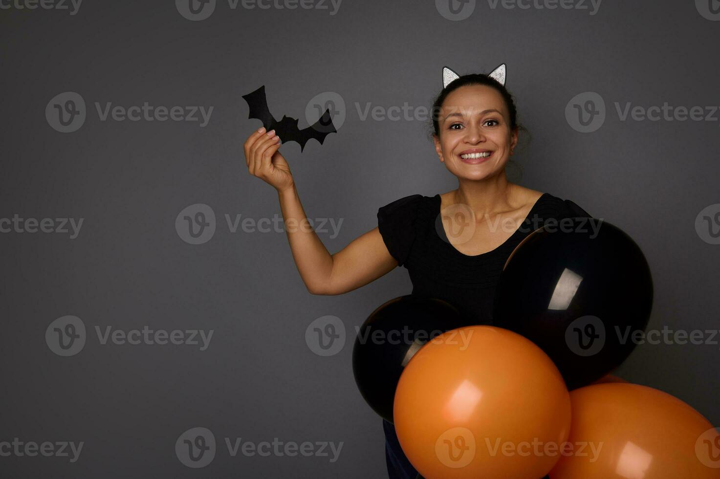 glad latinamerikan kvinna klädd i halloween karneval kostym utseende på kamera, innehar handgjort fladdermus i henne hand , Framställ med färgrik svart orange luft ballonger mot grå bakgrund med kopia Plats foto