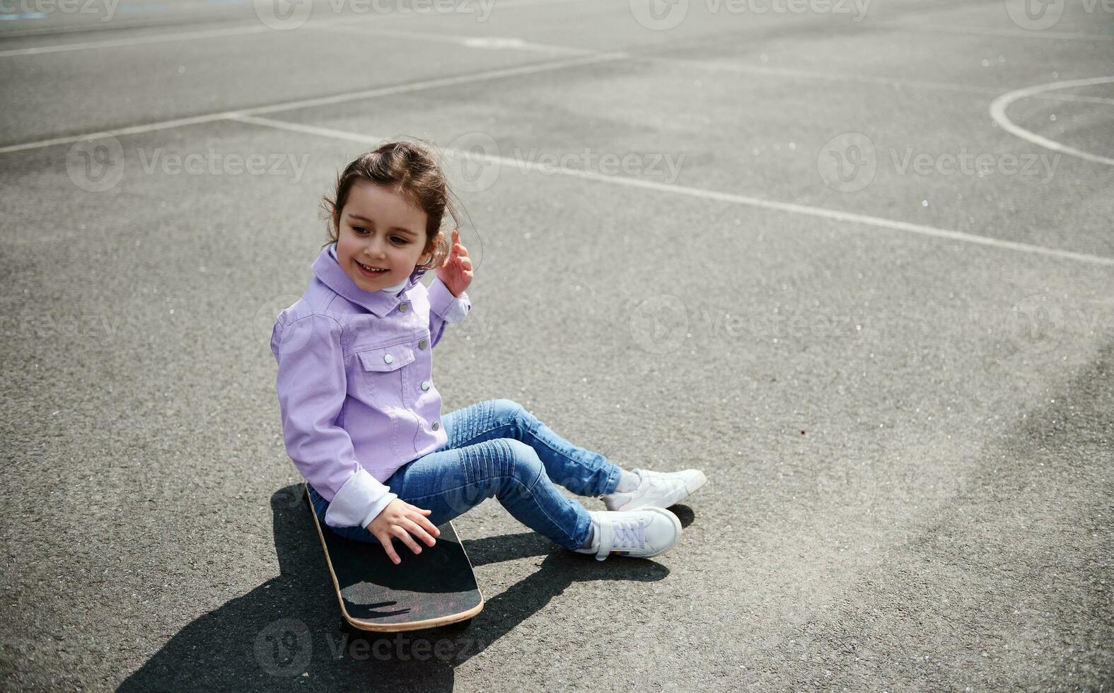 söt liten flicka Sammanträde på en skateboard på de lekplats på en skön solig dag foto