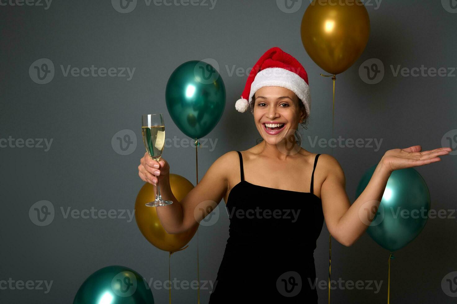 skön kvinna i santa hatt gläds och har roligt på jul fest innehar en champagne flöjt och kopia Plats på henne hand handflatan upp, isolerat på grå bakgrund med skinande guld och grön luft ballonger foto