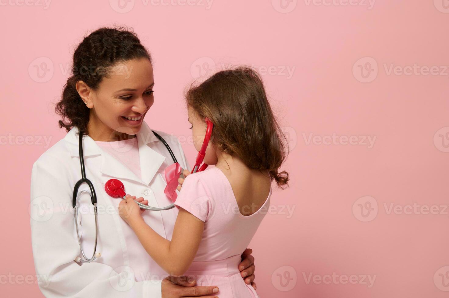 Söt flicka i rosa kläder användningar stetoskop, lyssnar till bröst och lungor andas av läkare med rosa band på bröst. begrepp av pedagogisk program för stödjande bröst cancer patienter. kopia Plats foto
