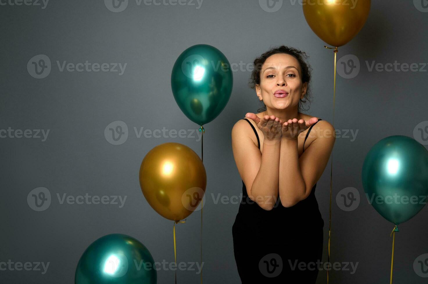 attraktiv latinamerikan kvinna klädd i elegant svart klänning står mot grå bakgrund med gyllene grön metallisk uppblåst ballonger, skicka luft kyssar ser på kamera. jul firande begrepp foto