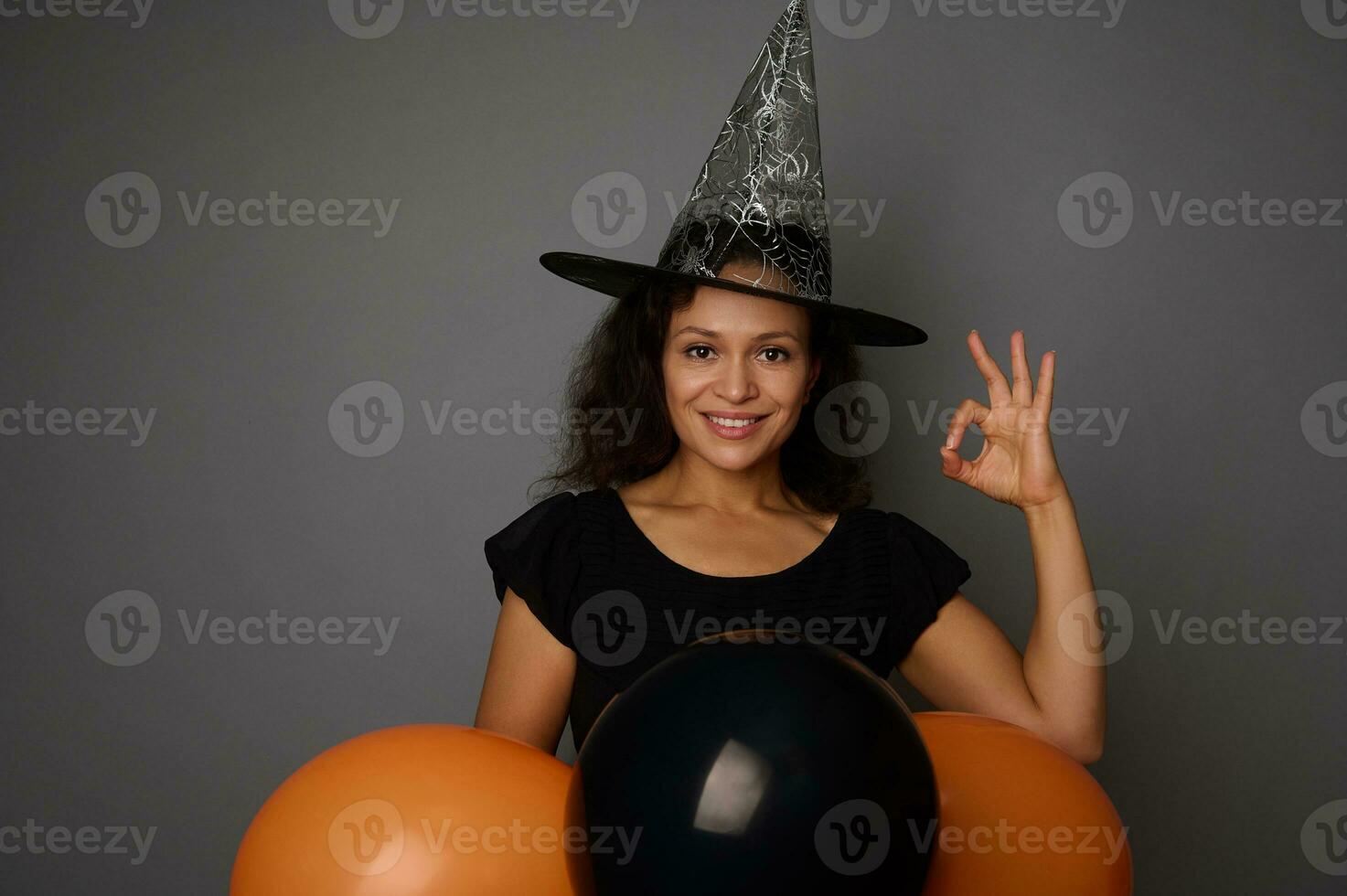 skön leende latinamerikan kvinna i trollkarl hatt, klädd i häxa karneval kostym för halloween fest, visar ok tecken, poser mot grå bakgrund med svart ballonger, kopia Plats foto