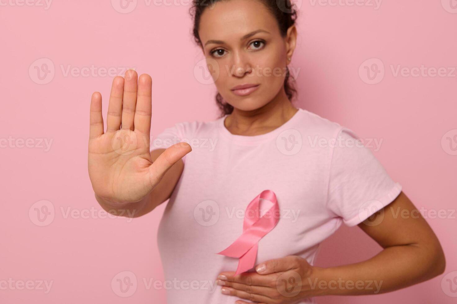 självsäker porträtt av suddig kvinna bär rosa t-shirt och cancer medvetenhet band. fokus på kvinnas hand gestikulerar sluta , isolerat över rosa bakgrund med kopia Plats foto