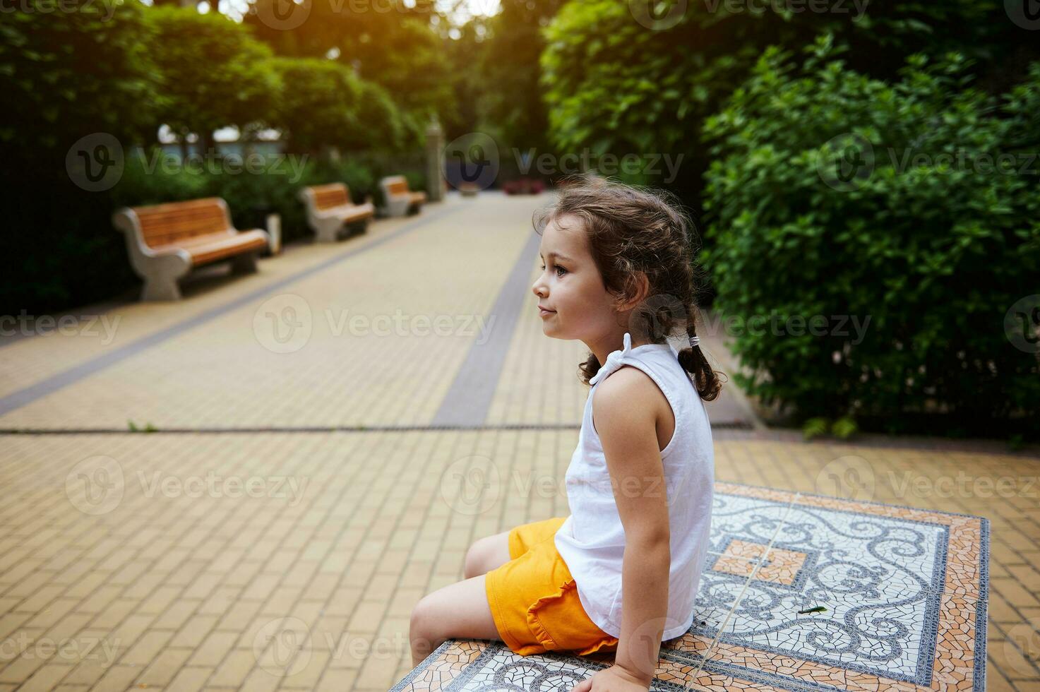 caucasian söt Lycklig liten barn flicka i ljus orange shorts och vit topp, Sammanträde på en bänk på de stad parkera gränd foto