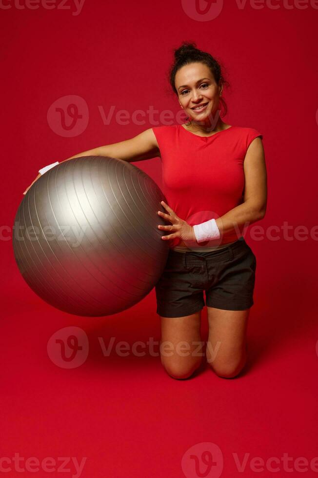charmig afrikansk amerikan atletisk kvinna övningar med passa boll, ler ser på kamera isolerat över röd bakgrund med kopia ad Plats foto
