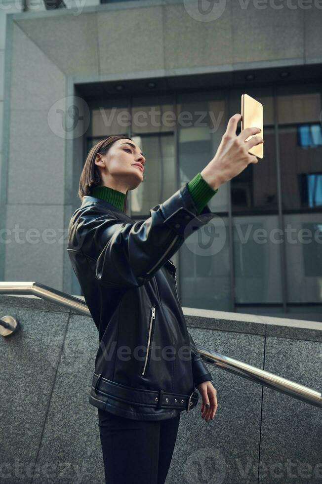 modern ung självsäker kvinna med eleganta kort frisyr bär svart eco läder jacka Framställ mot grå sten byggnad med en smartphone i henne utsträckt hand, tar selfie foto