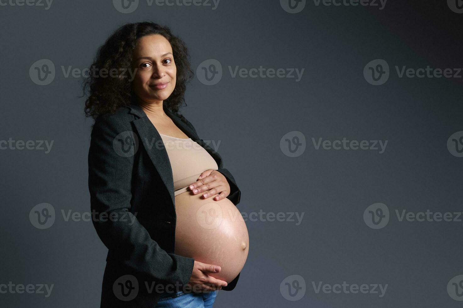 äkta leende Lycklig gravid kvinna innehav henne naken mage, ser på kamera, isolerat grå bakgrund. graviditet foto