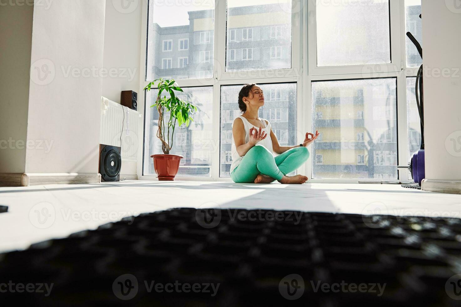 ung kvinna mediterar i de lotus placera mot de bakgrund av stor fönster i de Hem Gym. foto