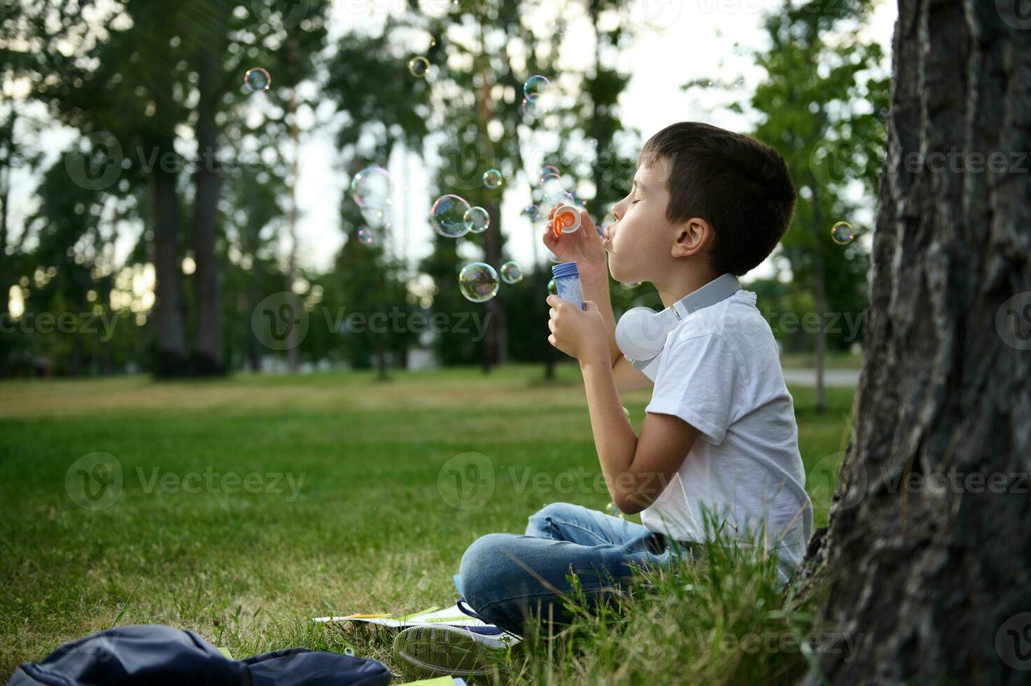 porträtt av ett förtjusande glad skön söt elementärt åldrig skola pojke njuter hans rekreation mellan klasser, blåser tvål bubblor, Sammanträde på de grön gräs av urban stad parkera foto