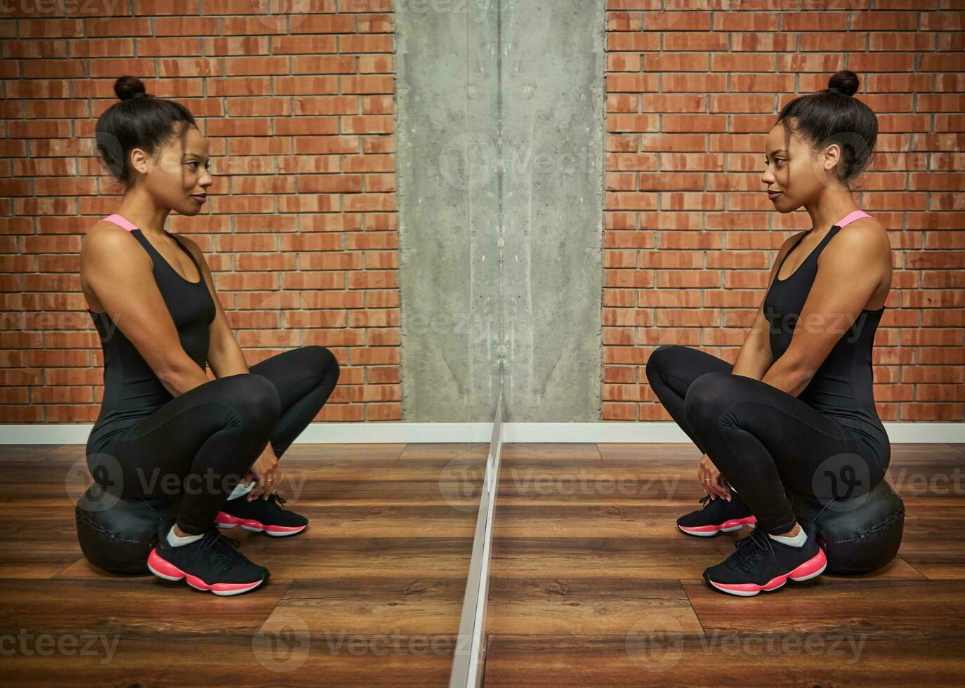 porträtt av en skön sportig idrottare afrikansk kvinna i svart sportkläder ser på henne spegel reflexion, Sammanträde på en medicin kondition boll mot röd tegelstenar vägg bakgrund av en Gym klass foto