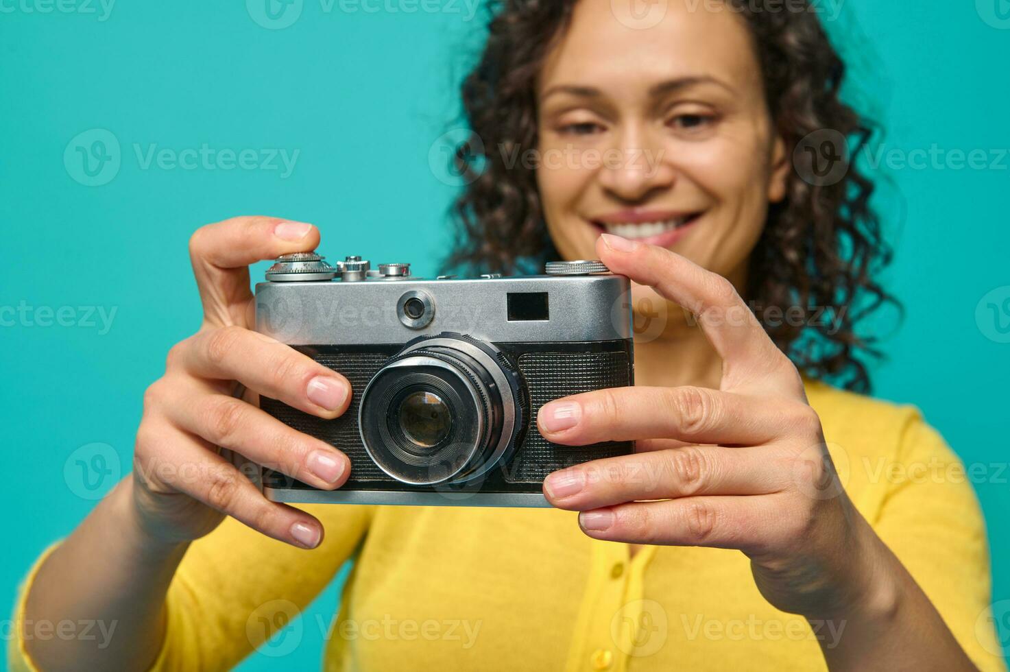 fokus på en årgång kamera- gammal retro stil fotografisk Utrustning i de händer av en suddig leende glad kvinna med lockigt hår, klädd i ljus gul Tröja, isolerat över blå bakgrund. foto